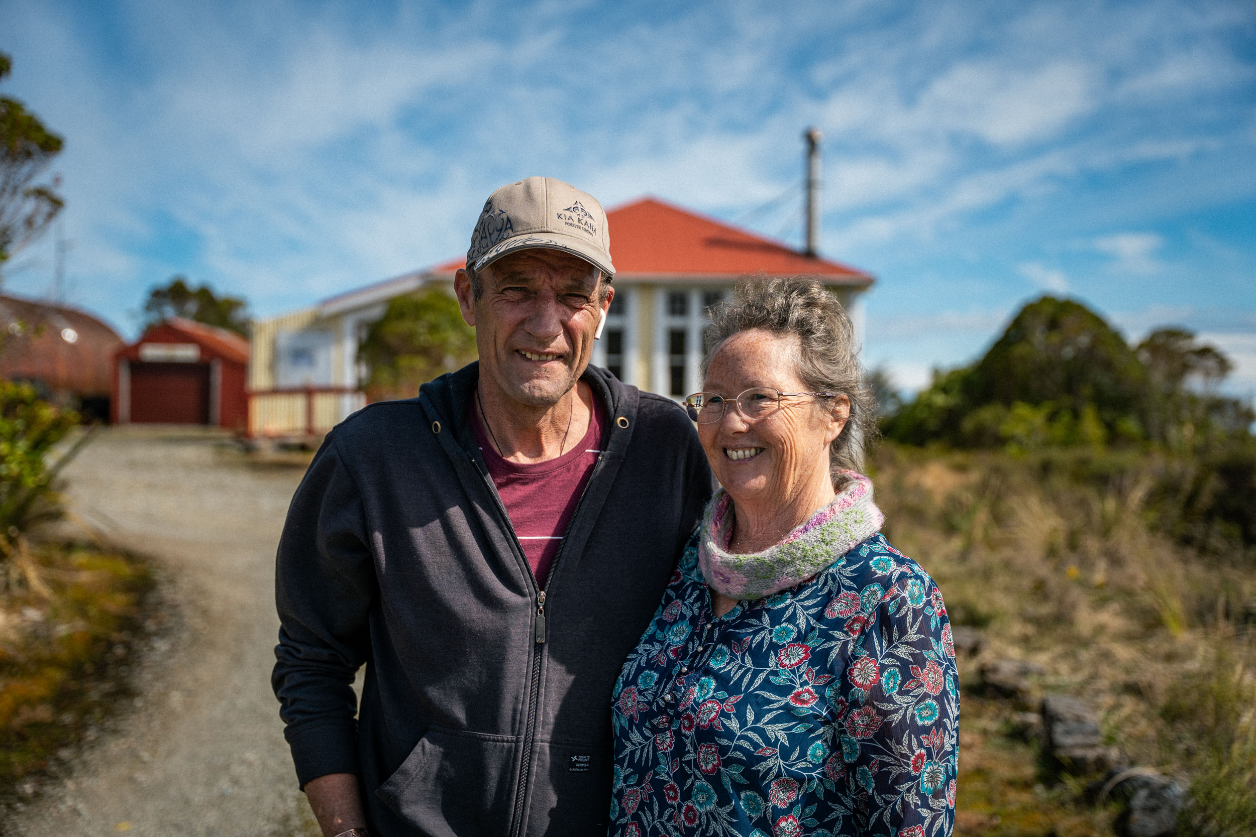 man and woman standing in front of a weatherboard house with a red roof and chimney, blue sky behind them
