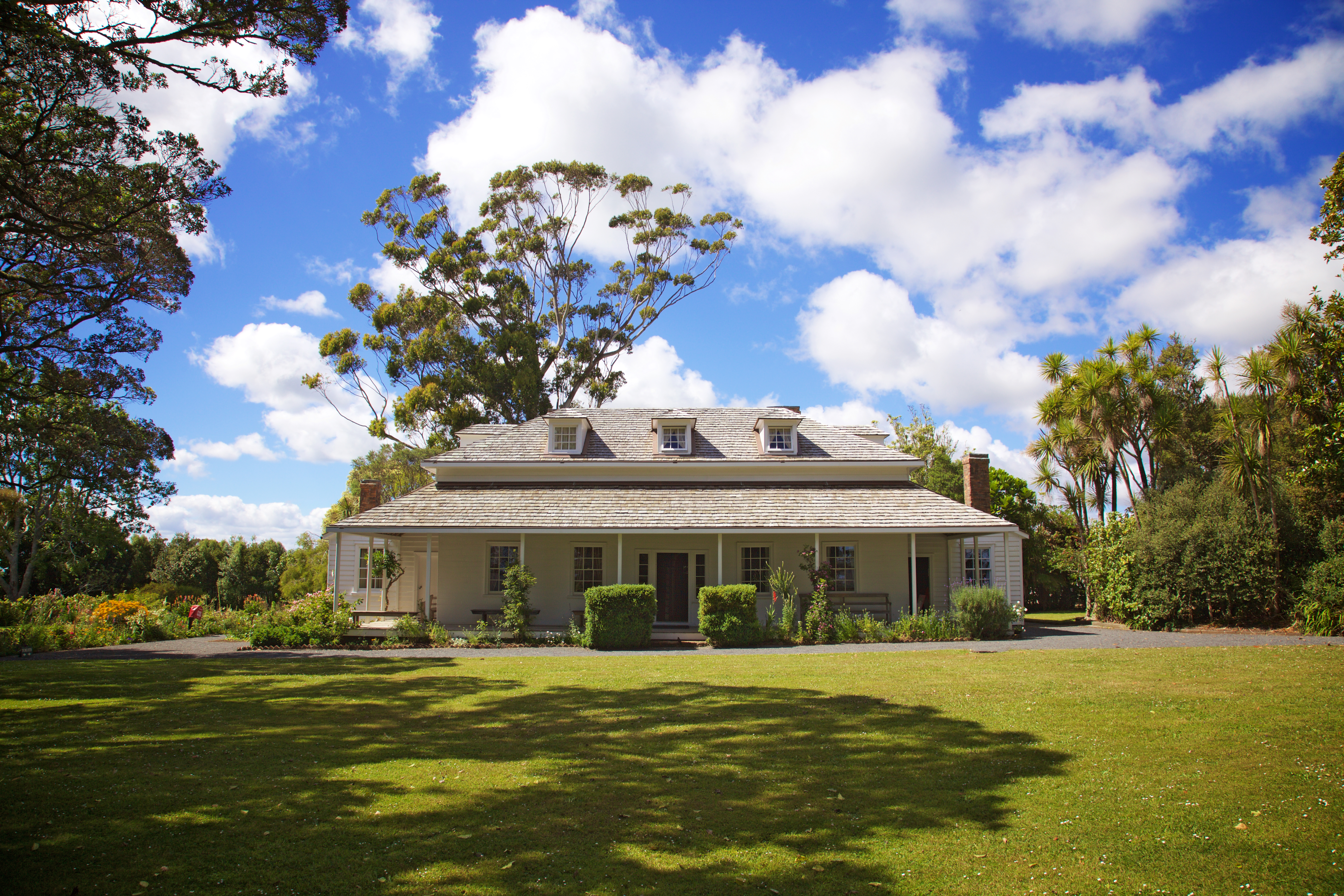colonial weatherboard building with shingled roof and verandah, with lawn and blue sky