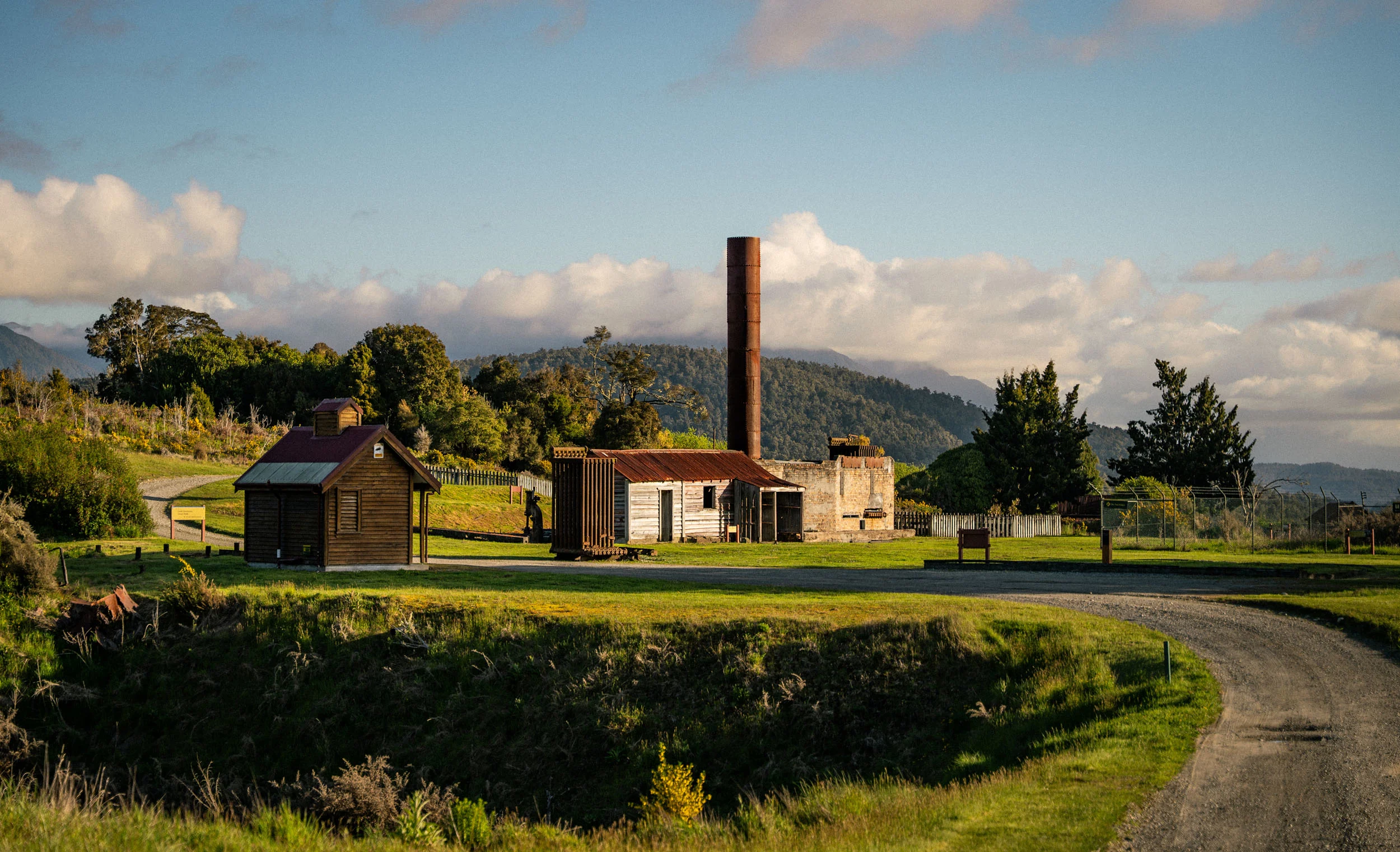 A curved road leading to old mining buildings one with a chimney, with rolling green hillside around. 