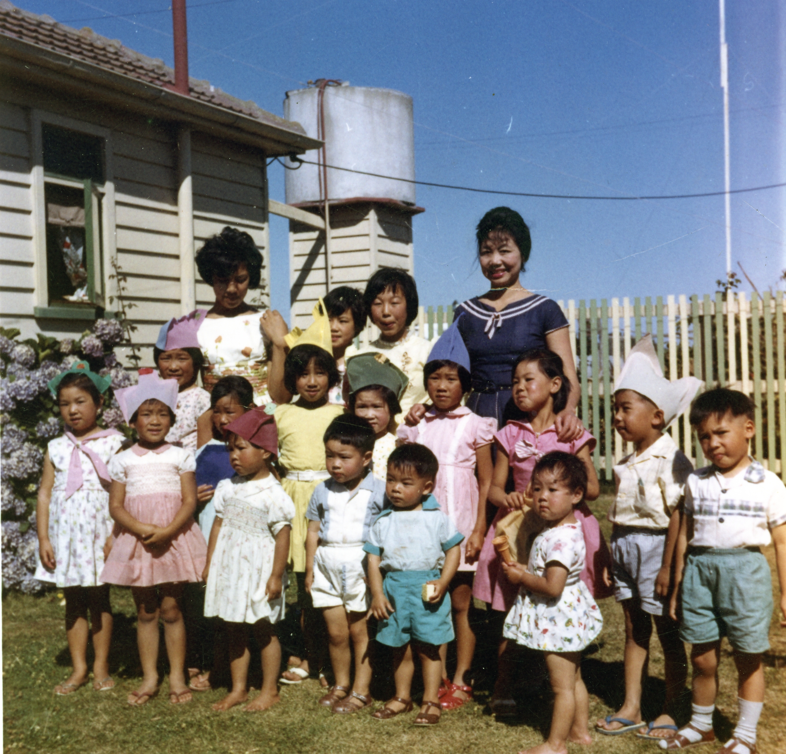 Group of Chinese children in colourful clothes and party hats, with several young Chinese women standing behind them.