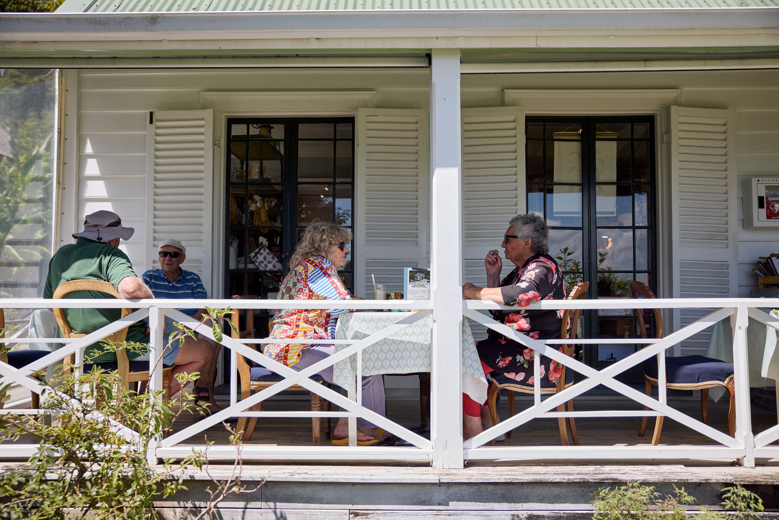 Two ladies sit at table on verandah of old white wooden house.