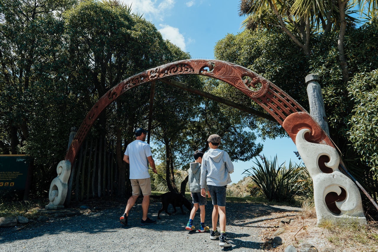man and two boys walk under arch with Māori designs, trees in background