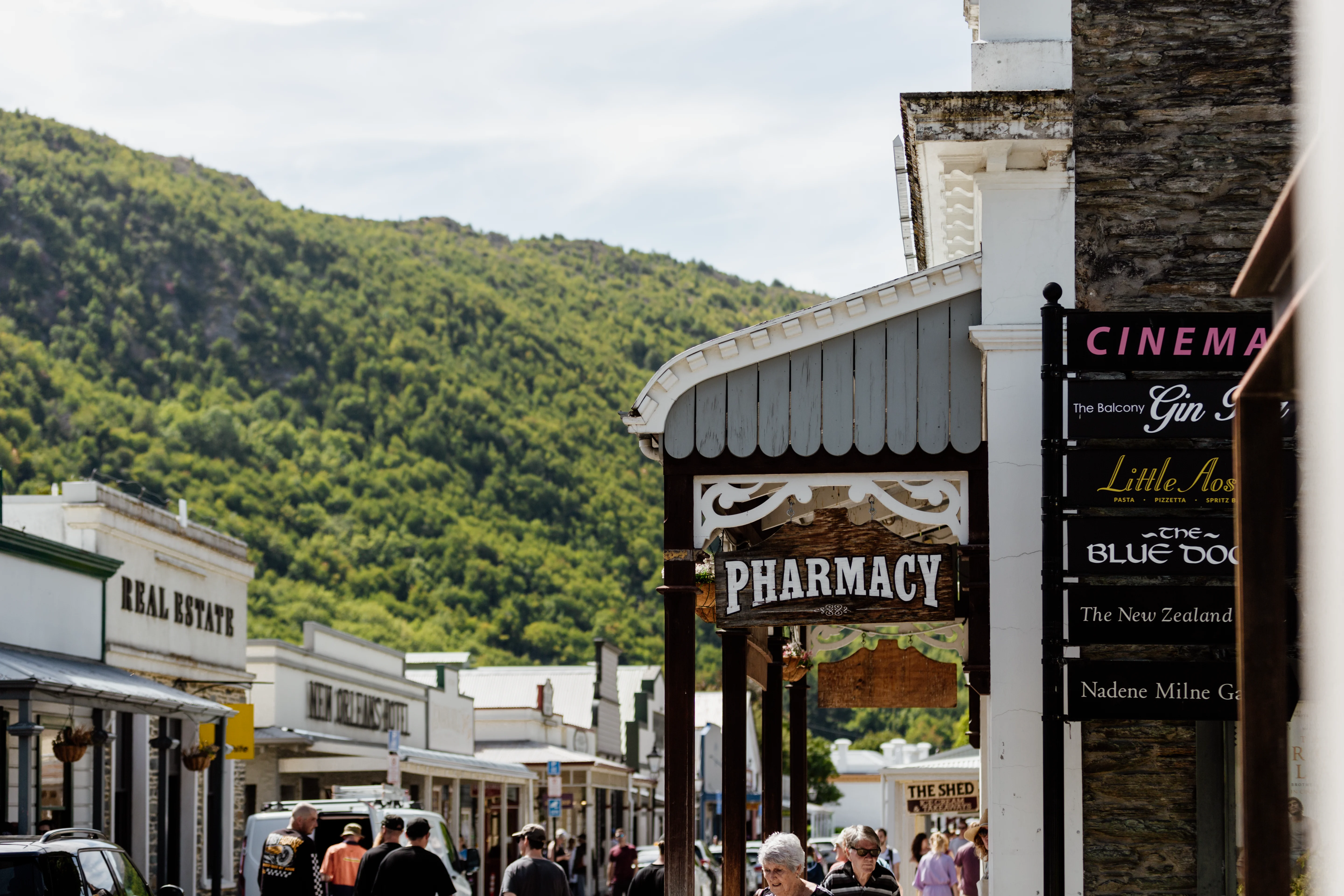 Bustling main street of Arrowtown with restored goldrush era shop fronts and a green forested hill behind. 