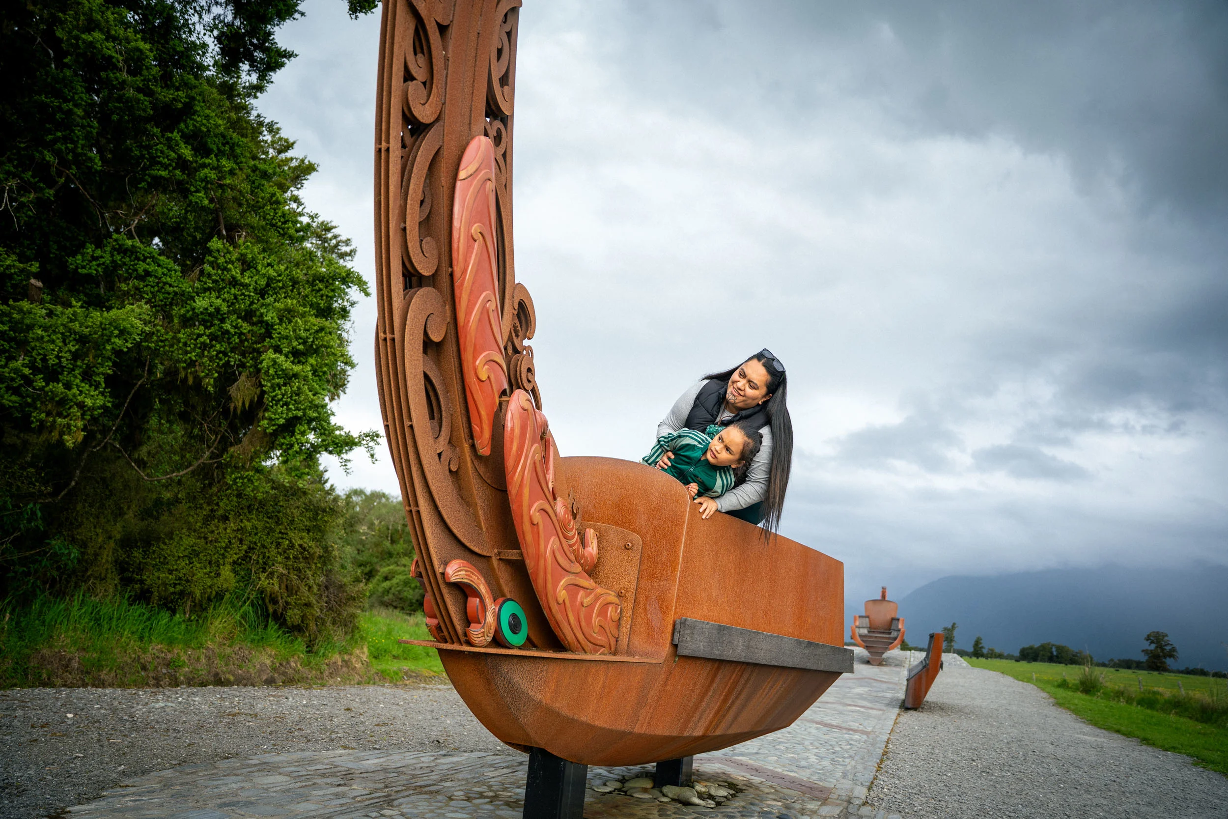 A young boy and a woman are aboard a sculpture of a waka, made in rusted corten steel with Māori carvings on. 