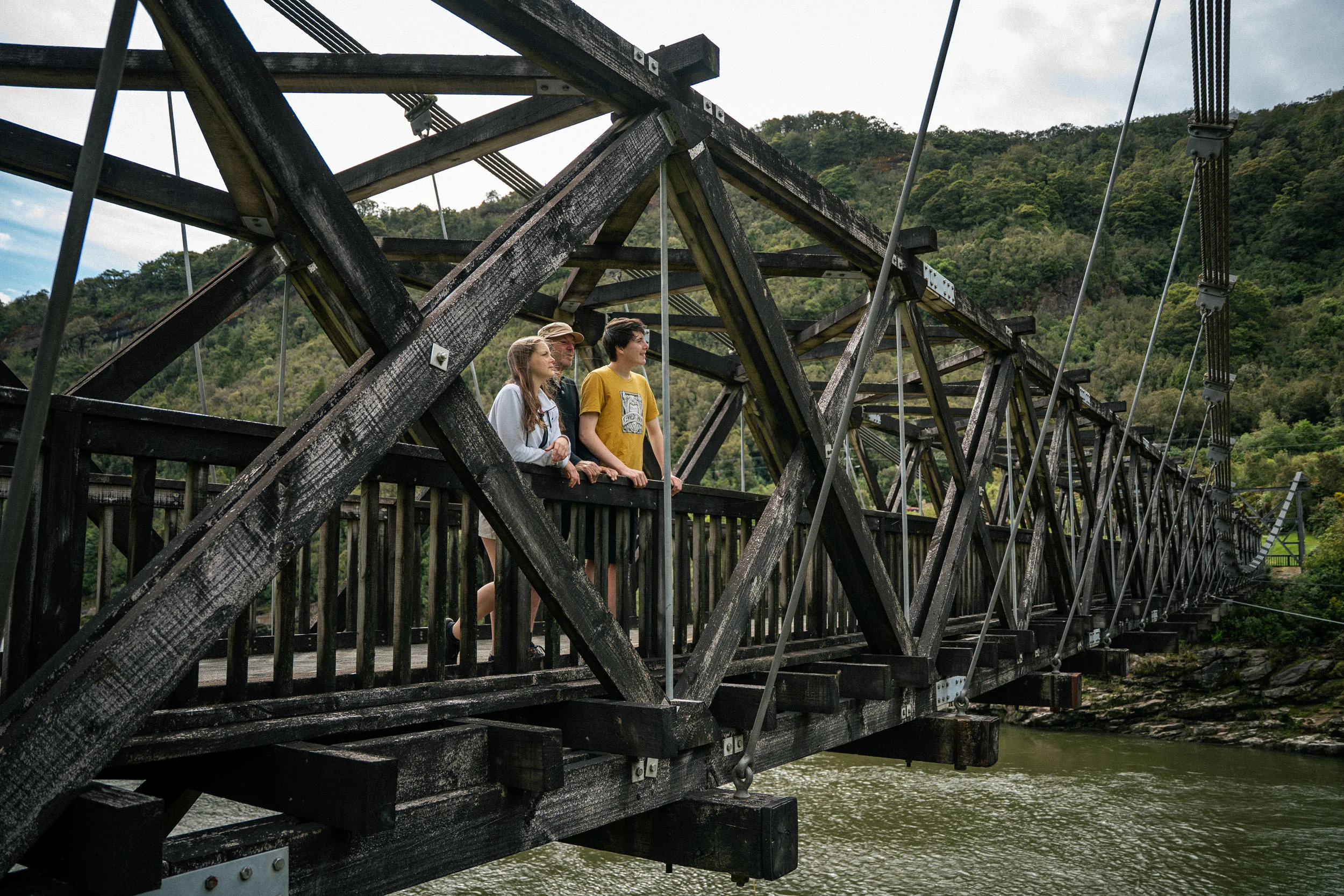 A teenage girl and boy look out standing on a large iron bridge. Steep green hills behind. 
