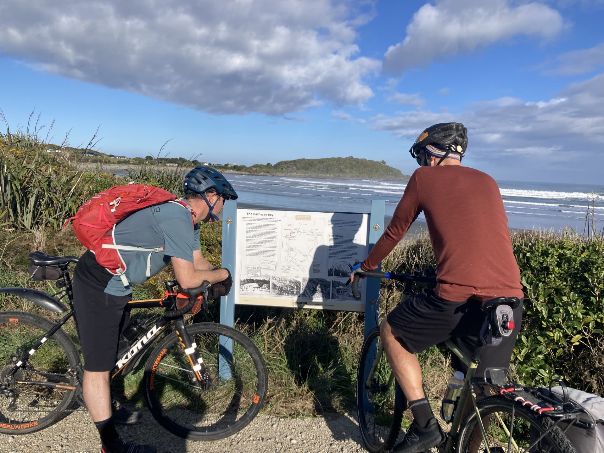 Two men on bikes reading an interpretation sign against coastal view