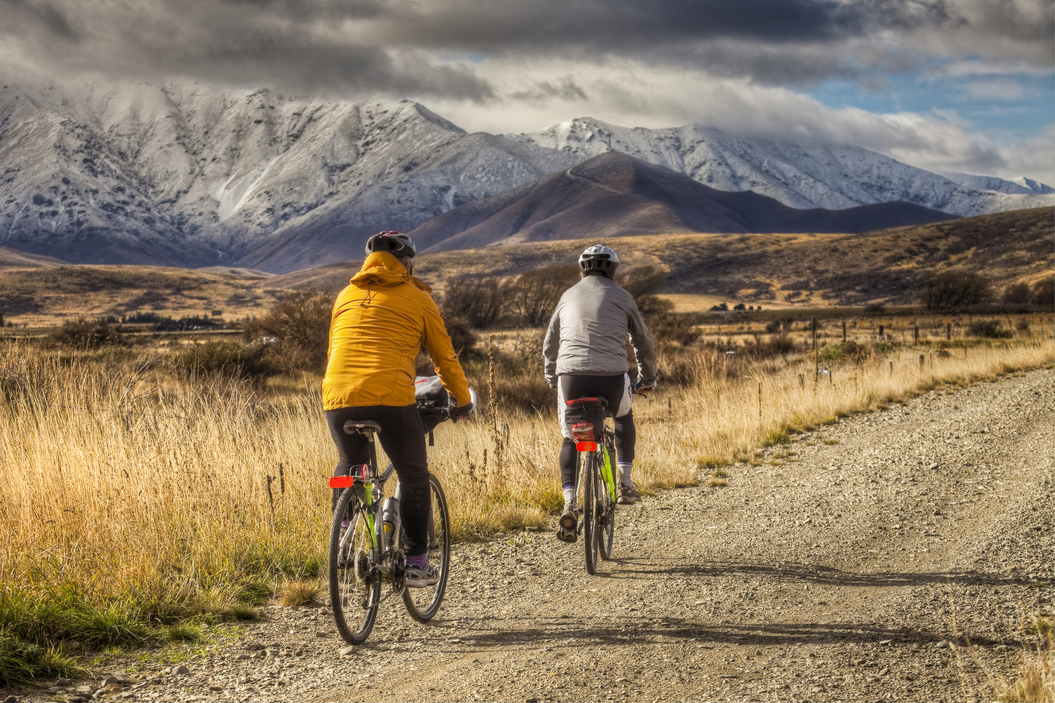 two people cycling in Otago landscape with mountains, one wearing yellow jacket