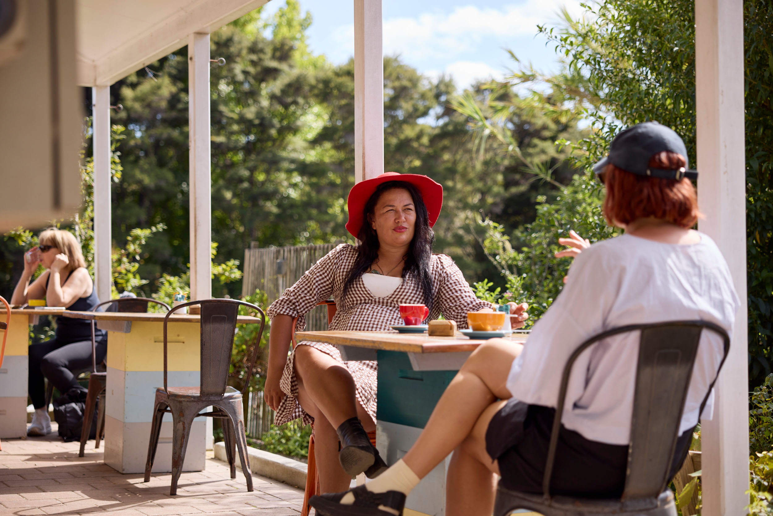 two woman sit at table on verandah, with coffees, trees in background