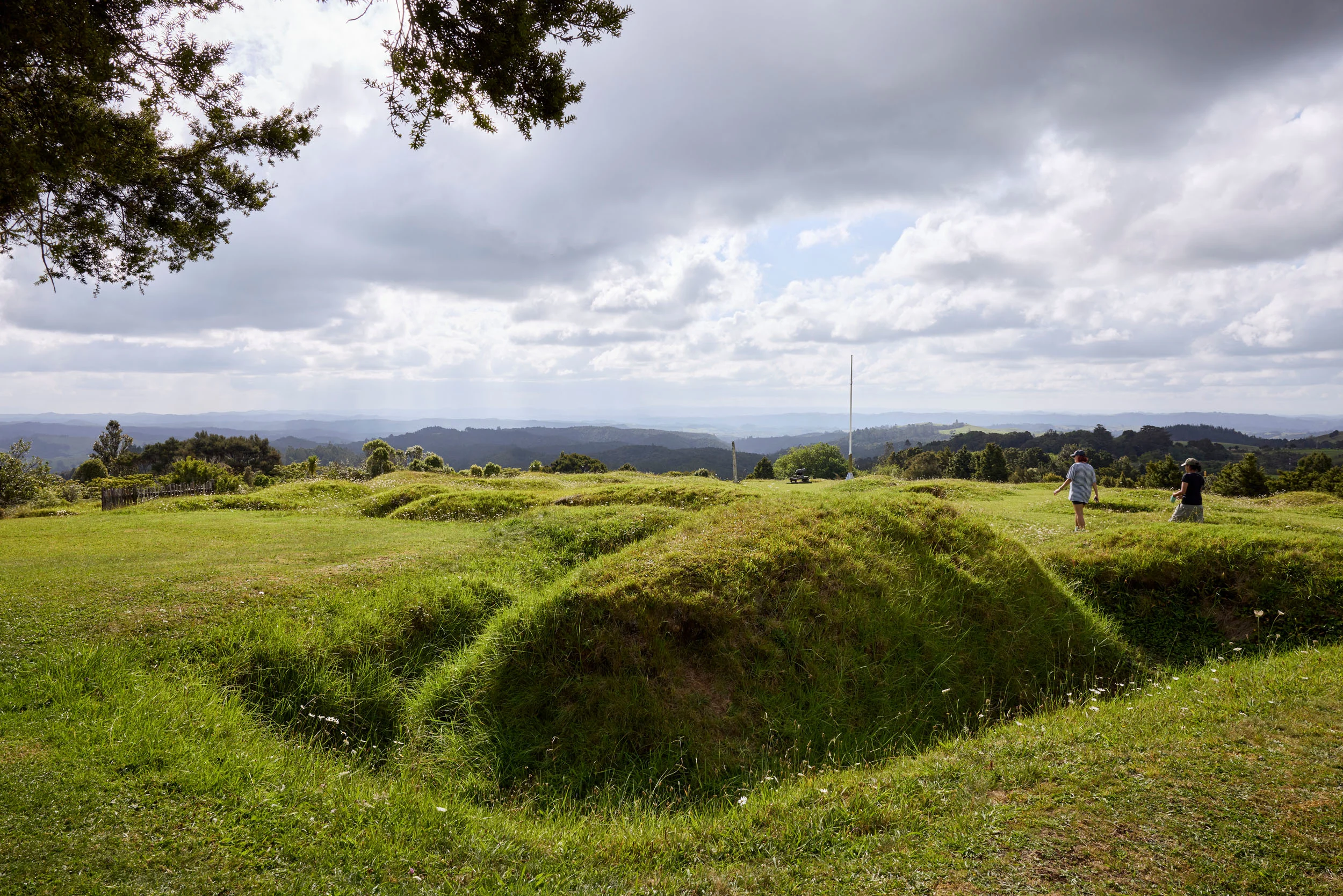 A wide shot of the grassy pā area with deep trenches all around, two people walking away. A view off into the distance of hills and sky. 