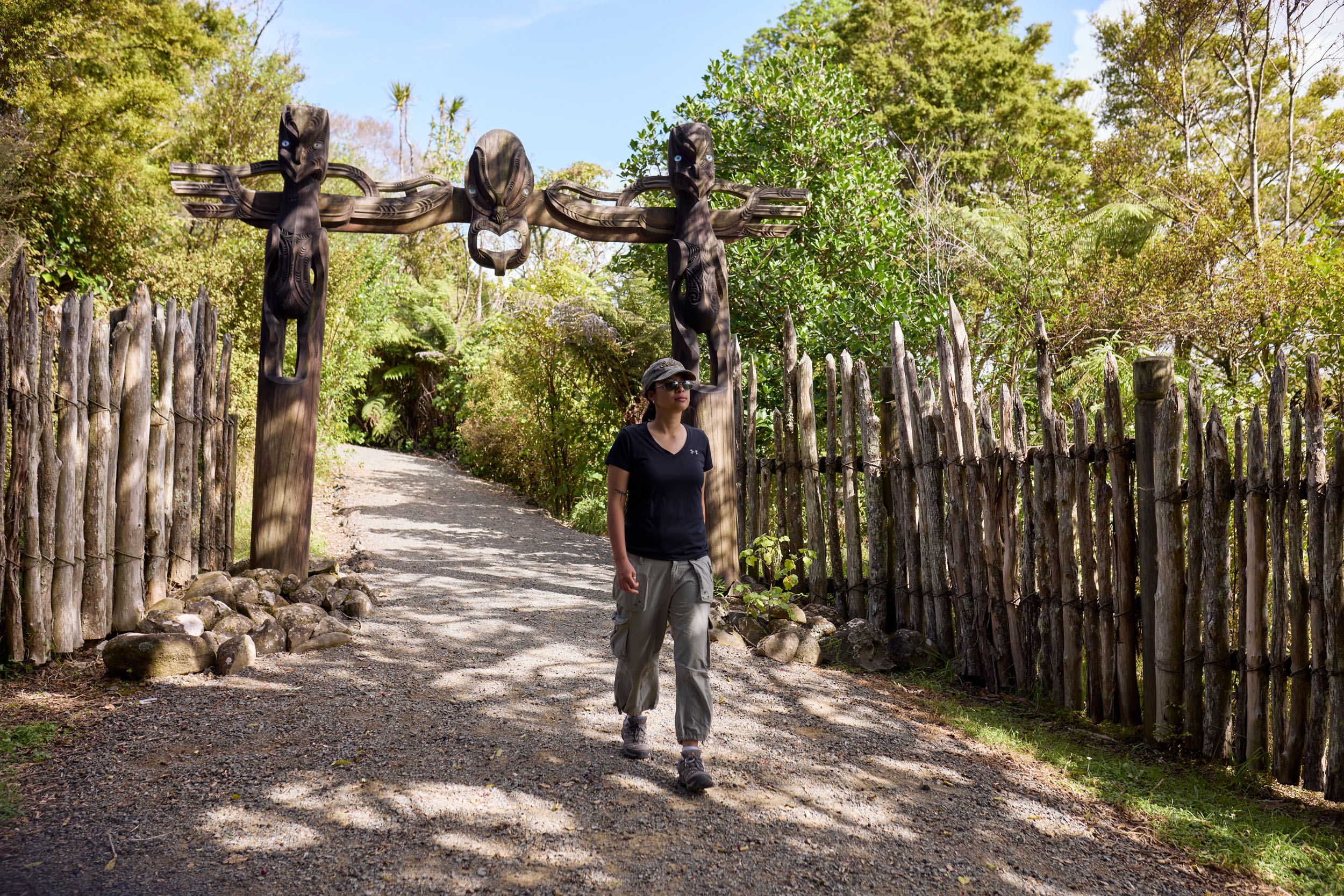 young woman walks between wooden palisade fencing and wooden arch, trees in background