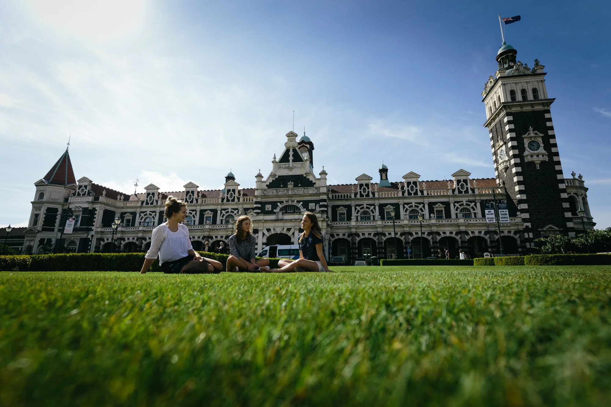 A wide shot of the grand Dunedin Railway Station with three young women in foreground on the lawn. 