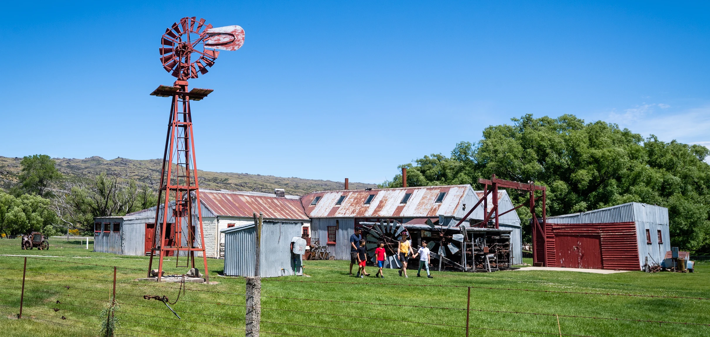 A man, woman and three children walk across a yard with an old windmill to the left and old corrugated iron sheds behind.