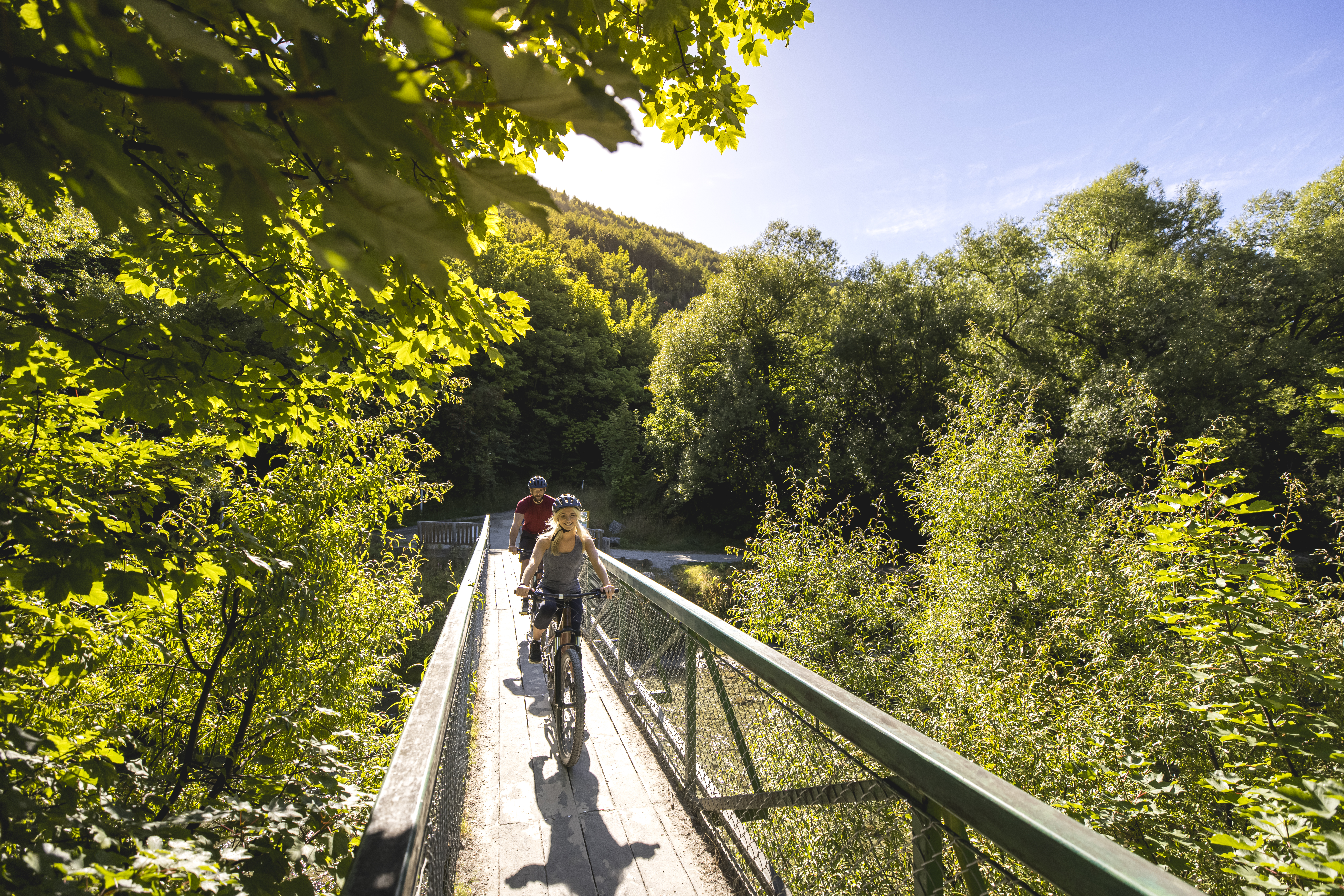 Cyclists biking across a wooden bridge over river, surrounded by trees