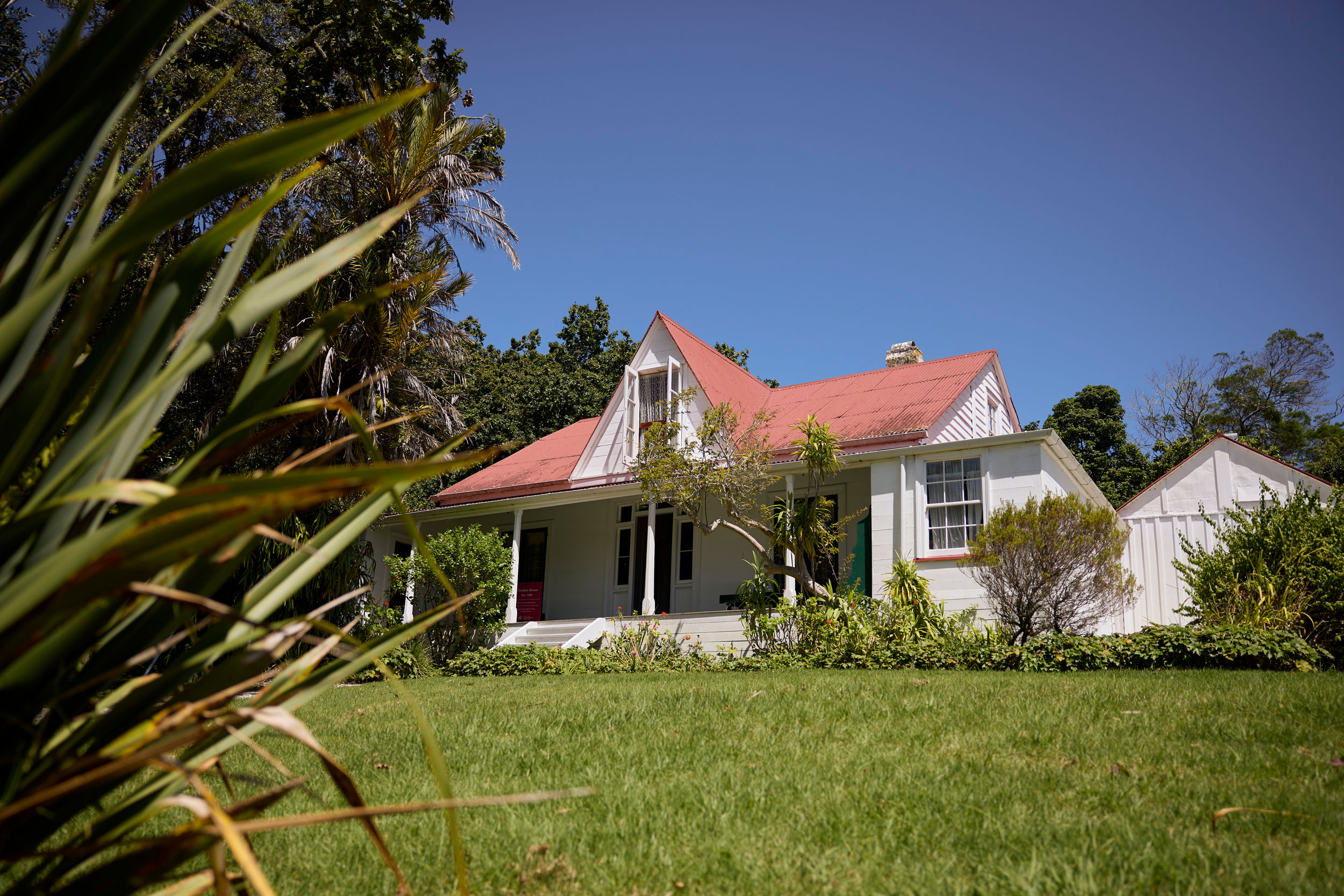 white weatherboard colonial house with red roof against blue sky, lawn and flax in foreground