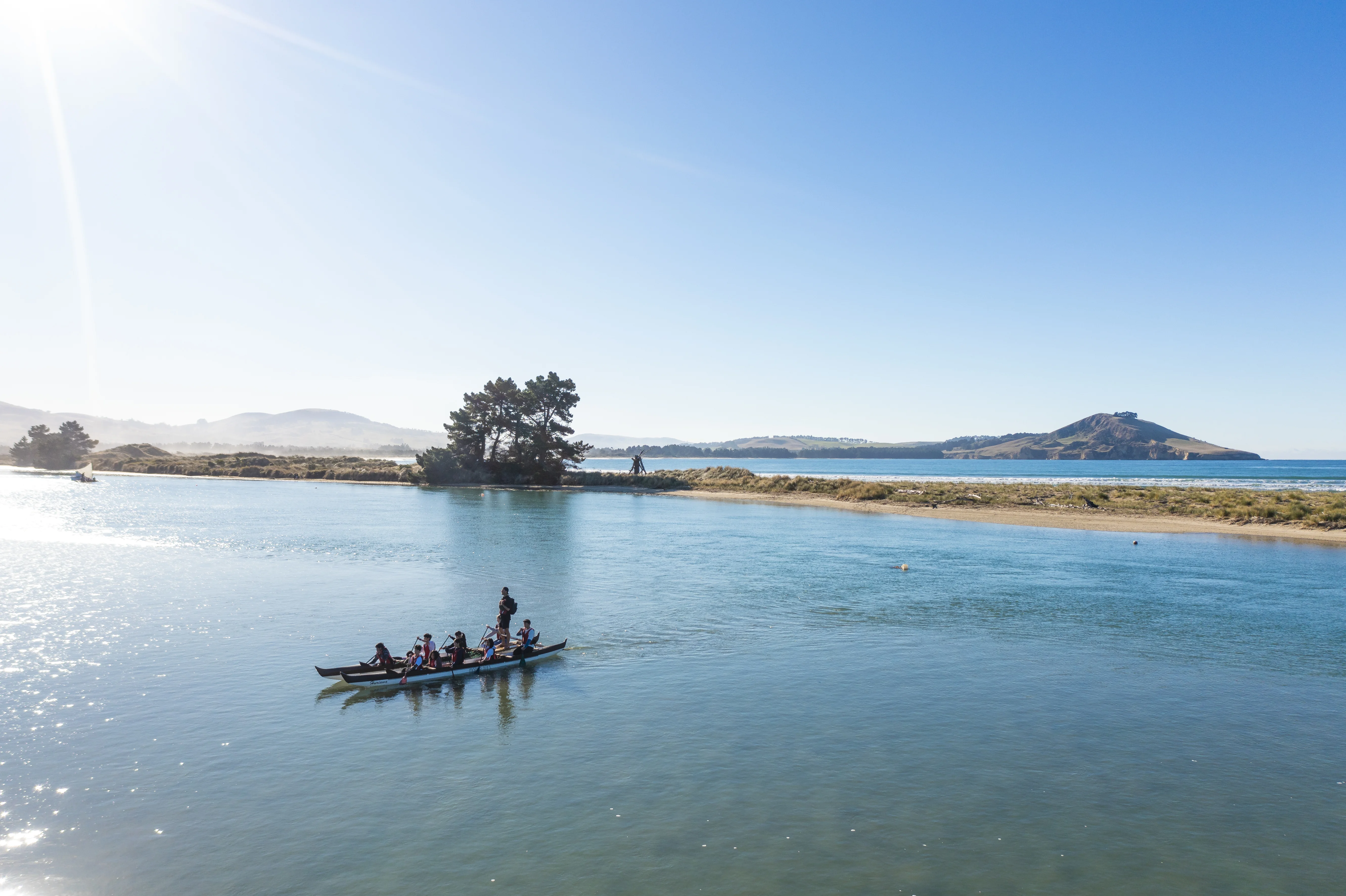 Wide shot of a sandy peninsula surrounded by turquoise water. A waka with people paddling in the foreground. 