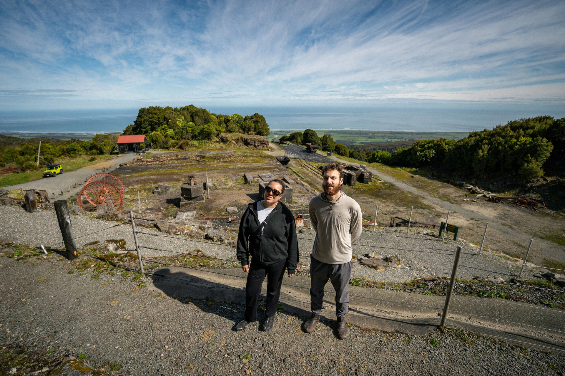 Two young people look up at the camera with vast mining site behind them, up high with the ocean in the distance. 