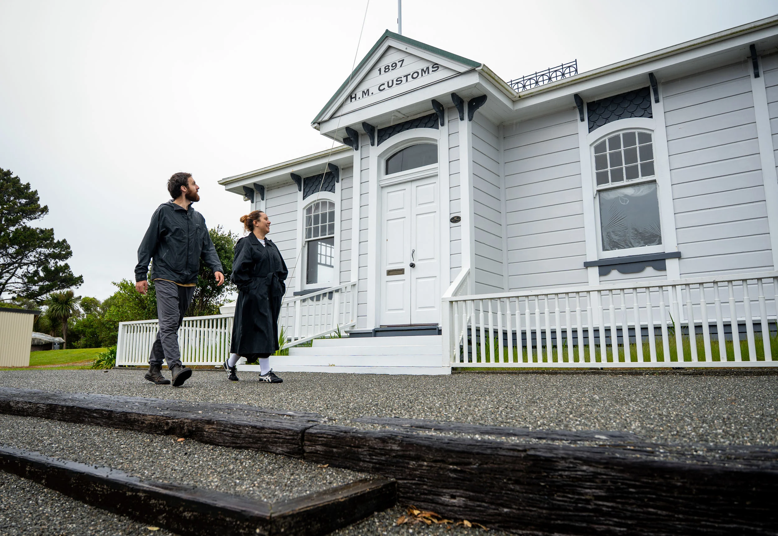 A Māori woman and Pākeha man wearing jackets walk past an historic customs building with 1897 on a sign. 