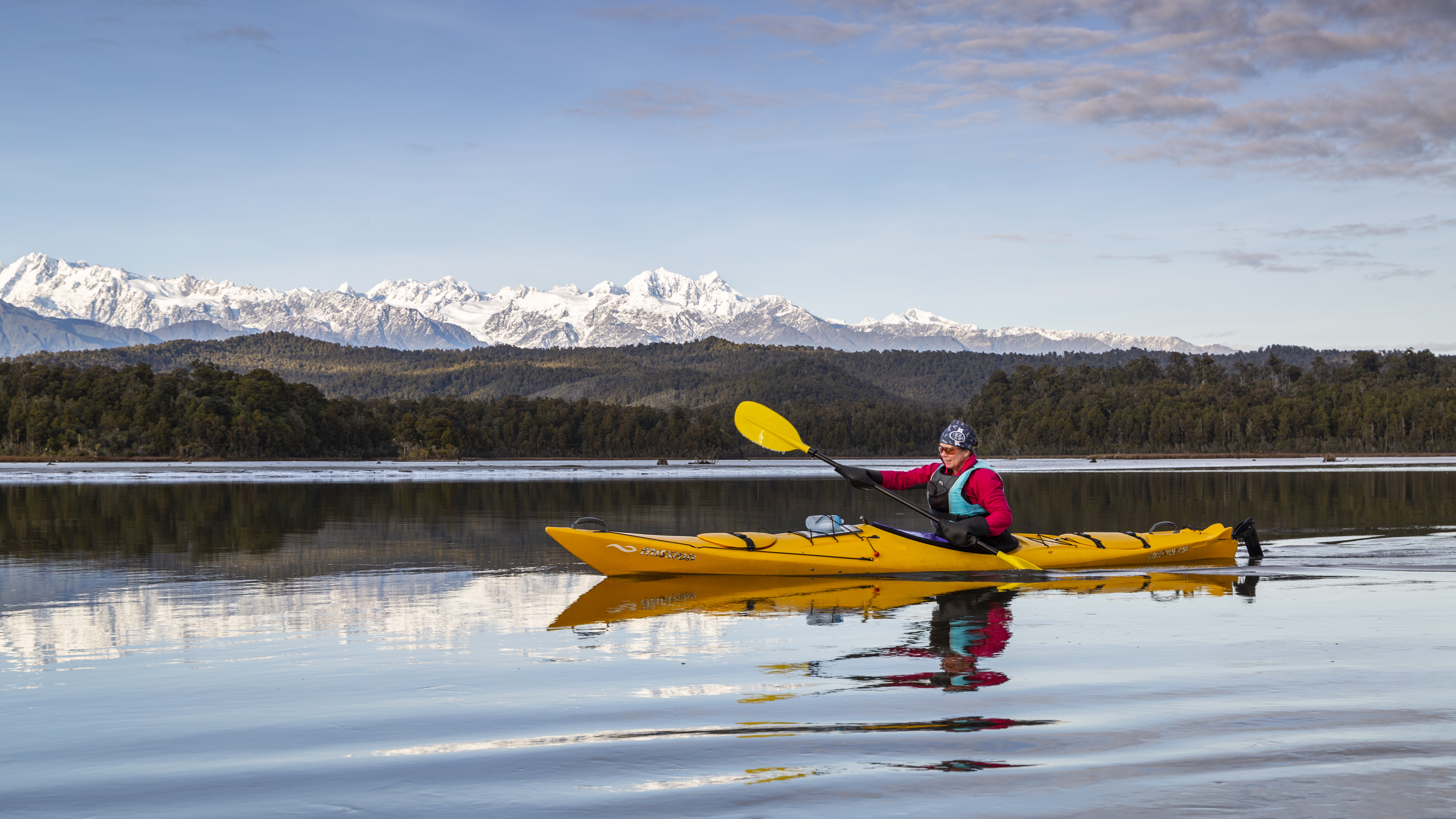 man in red jacket paddles in yellow kayak on still lagoon, snowy mountains in background