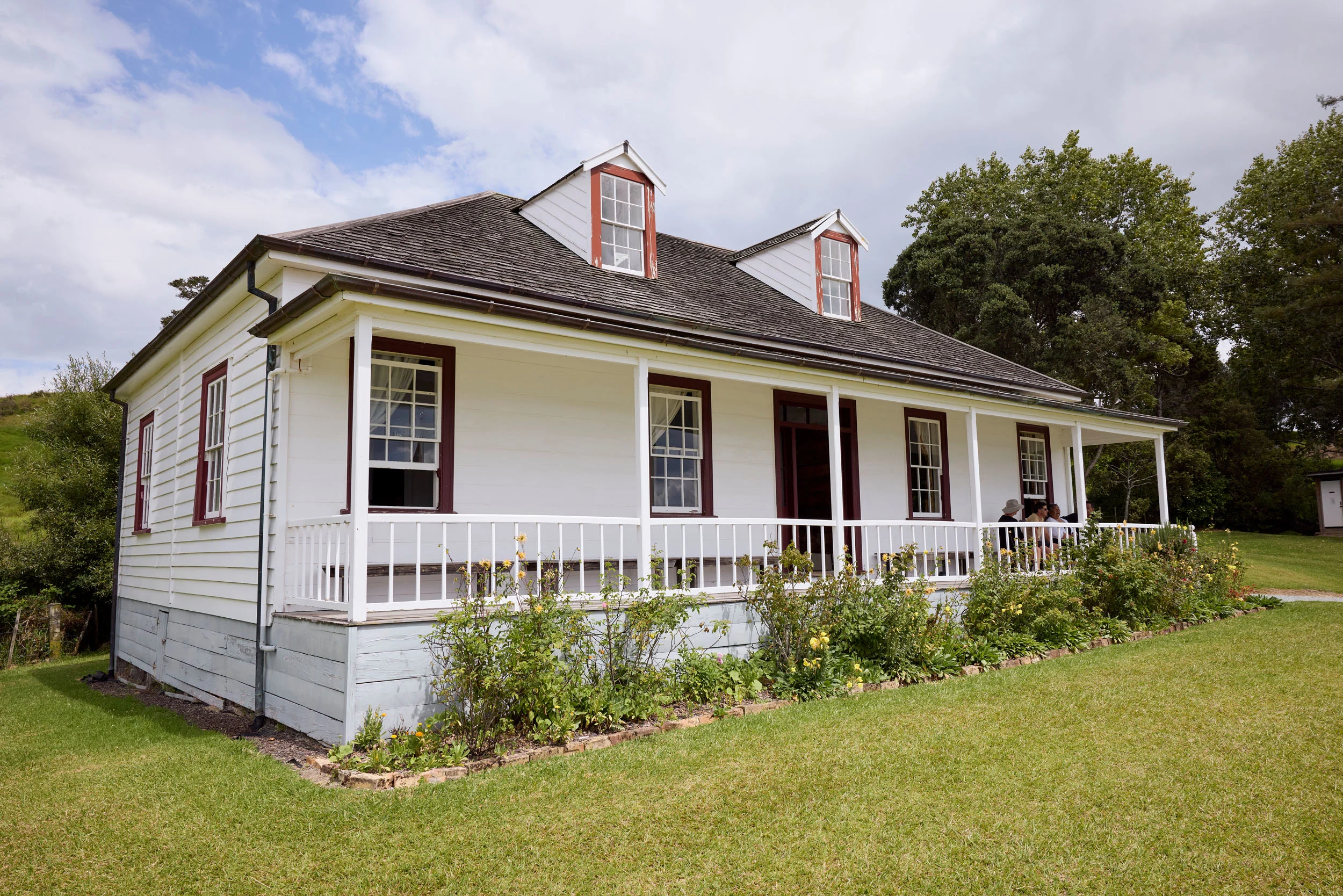 Old white mission house with grey slate roof on a green hill with flag flying in front. Four people sitting on veranda. 