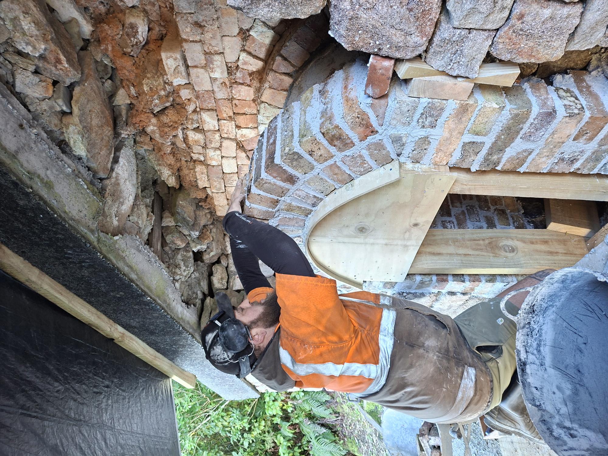 Man in orange jacket, black cap, working on brick arch