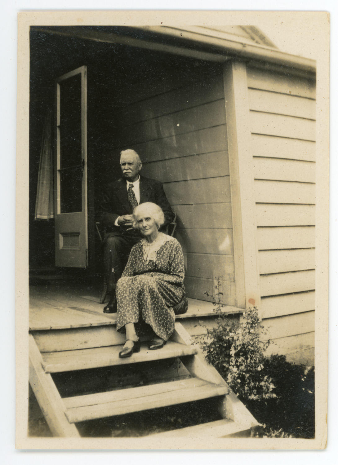 black and white photo of older man and women sitting on verandah of house above steps