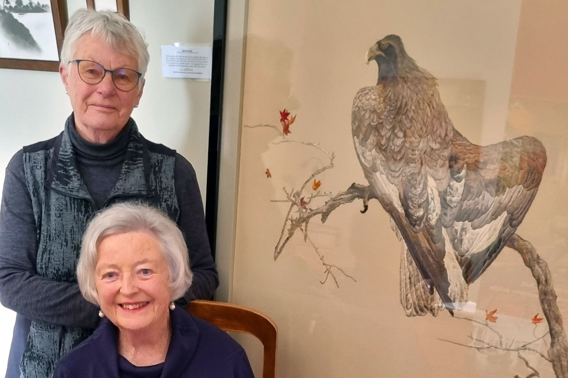 Two older white women with large framed embroidery of an eagle. 