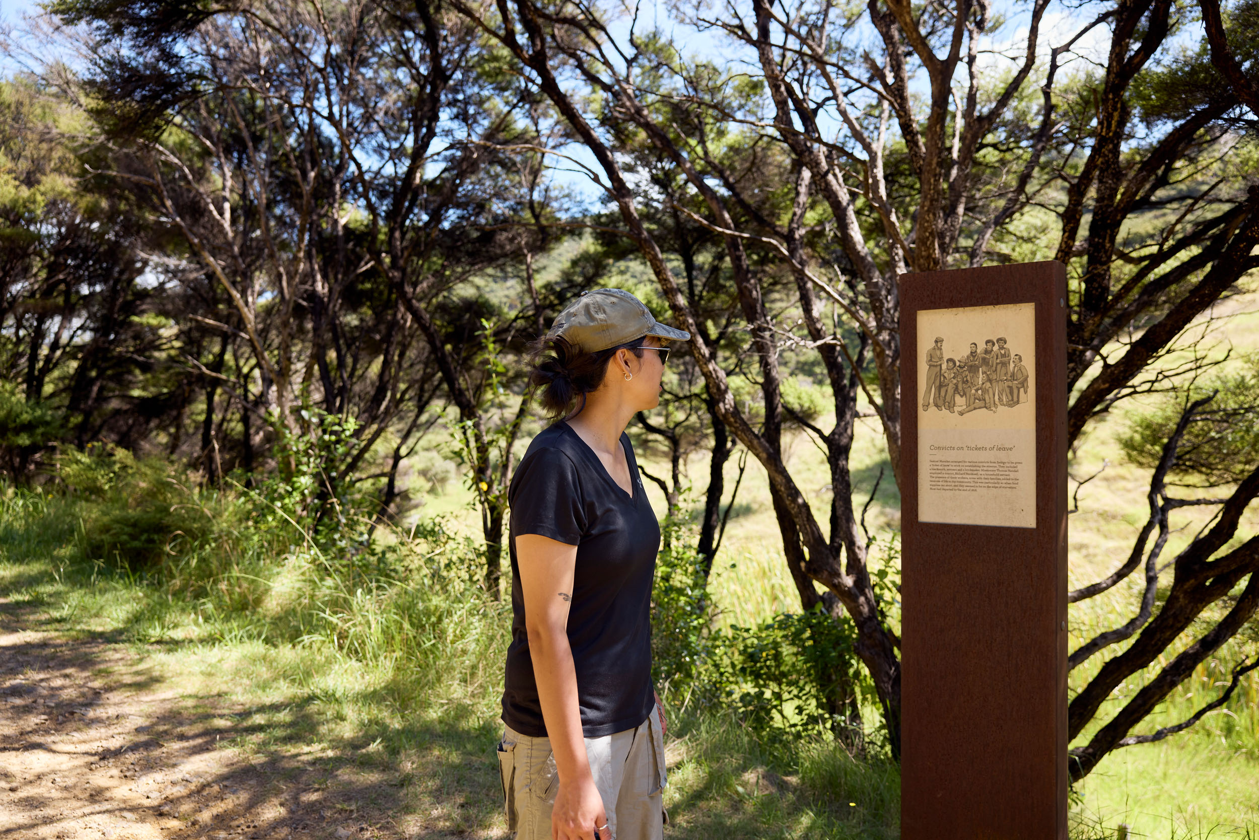 woman with cap and black t-shirt looking at metal interpretation sign with trees in background