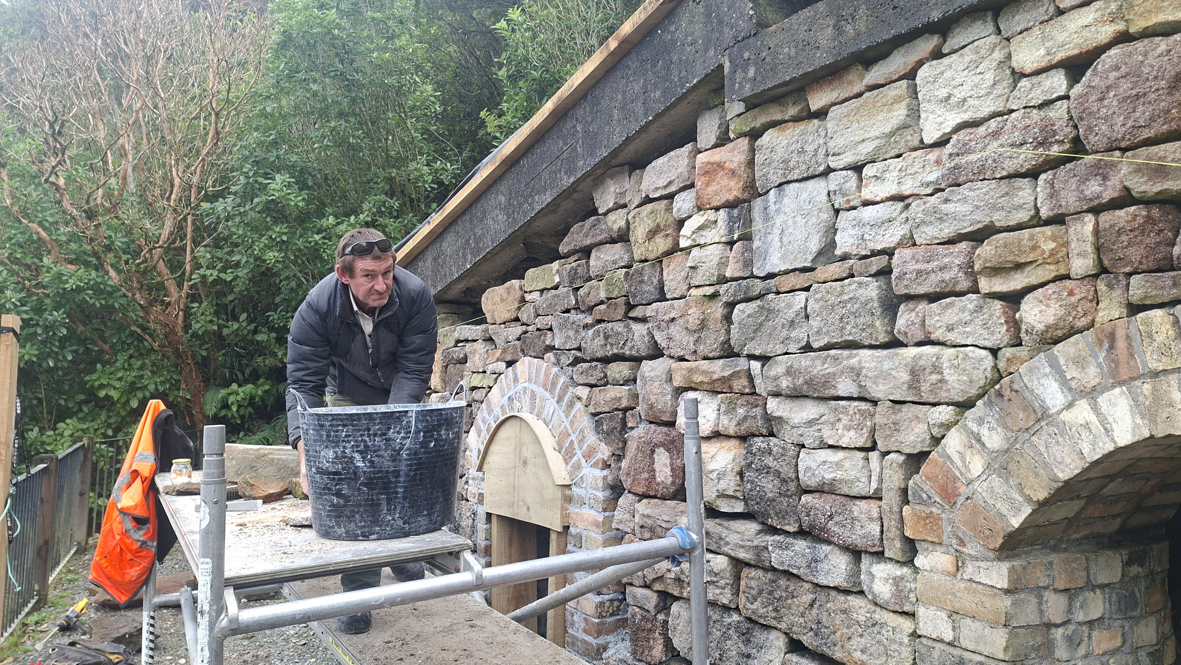 Man in black jacket mixing mortar in black bucket next to brick building with arches