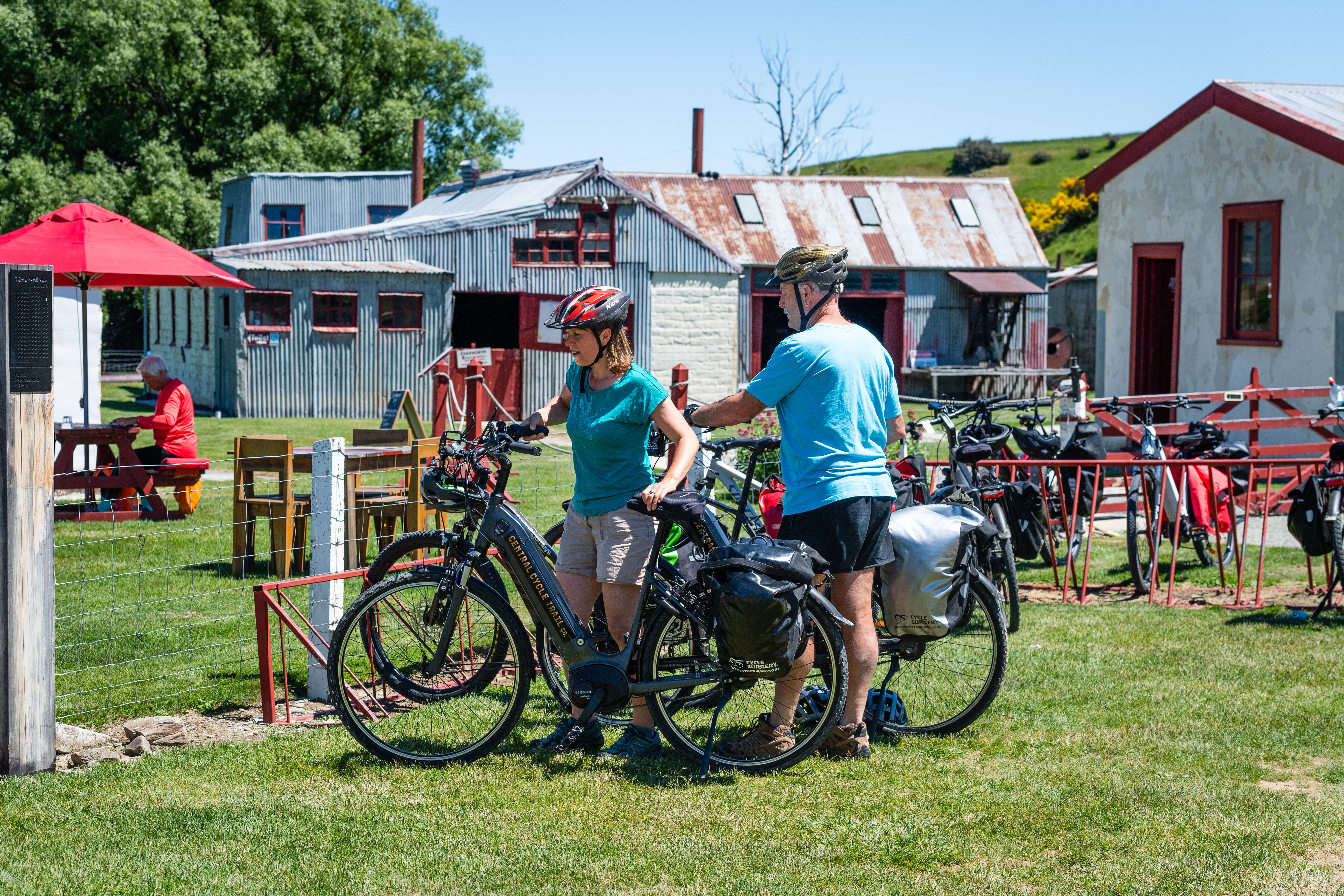 Cyclists with bikes next to fence, picnic tables and old buildings in background